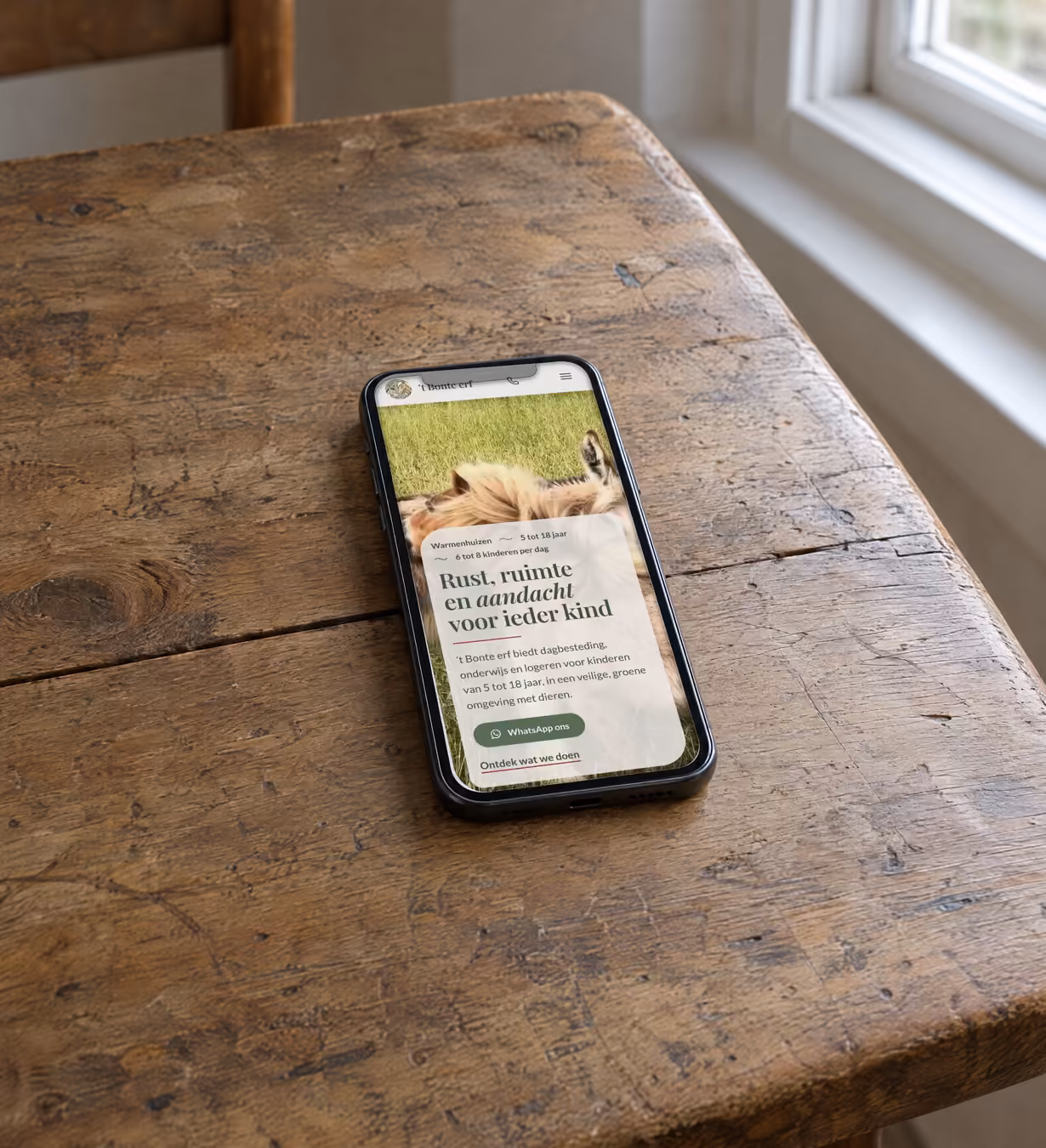 Telefoon schuin op een houten boerentafel bij een raam. Op het scherm de nieuwe website van 't Bonte erf: een ezel in het gras en een lichte tekstkaart met de kop "Rust, ruimte en aandacht voor ieder kind"