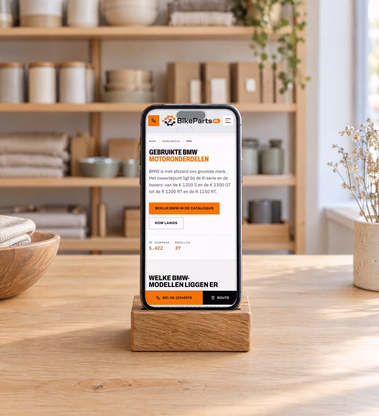 Telefoon in een eiken standaard op een lichte toonbank, overdag. Op het scherm de pagina over gebruikte BMW-motoronderdelen van BikePartsNL: een zwarte kop met een oranje regel, een oranje knop naar de catalogus en een lijst met BMW-modellen