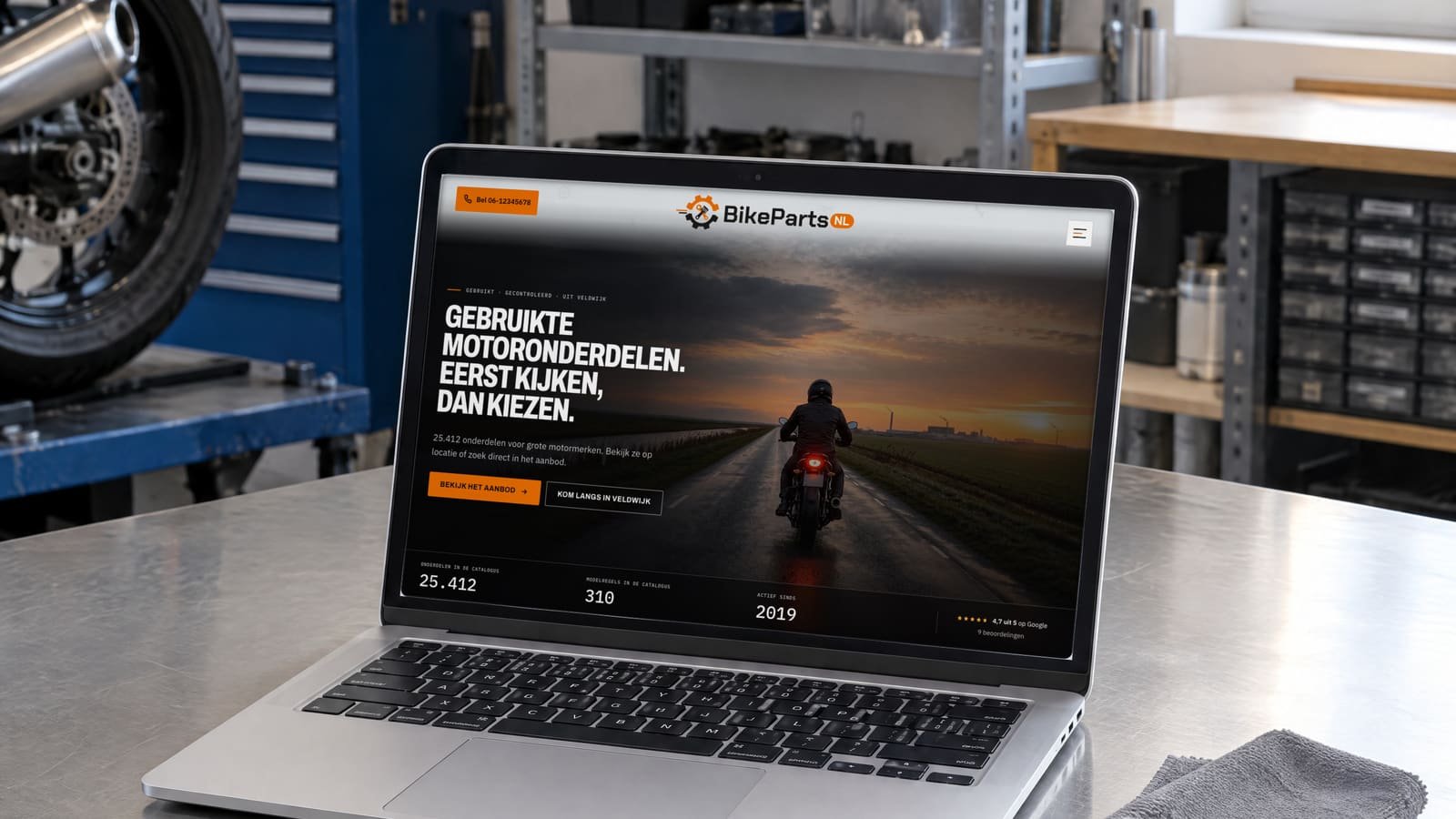 Een laptop op een stalen werkbank in een lichte motorwerkplaats, met op het scherm de website van BikePartsNL: de kop "Gebruikte motoronderdelen. Eerst kijken, dan kiezen." naast een motorrijder op een polderweg.