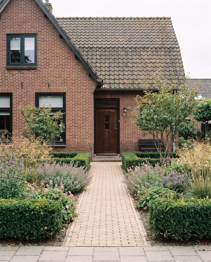 De voortuin van een bakstenen huis met een recht pad naar de voordeur, lage hagen en vaste planten.